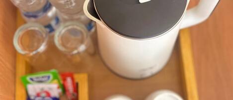 A white electric kettle with a black lid, two clear plastic bottles with blue caps, and a wooden tray with packets of tea and two white teacups.