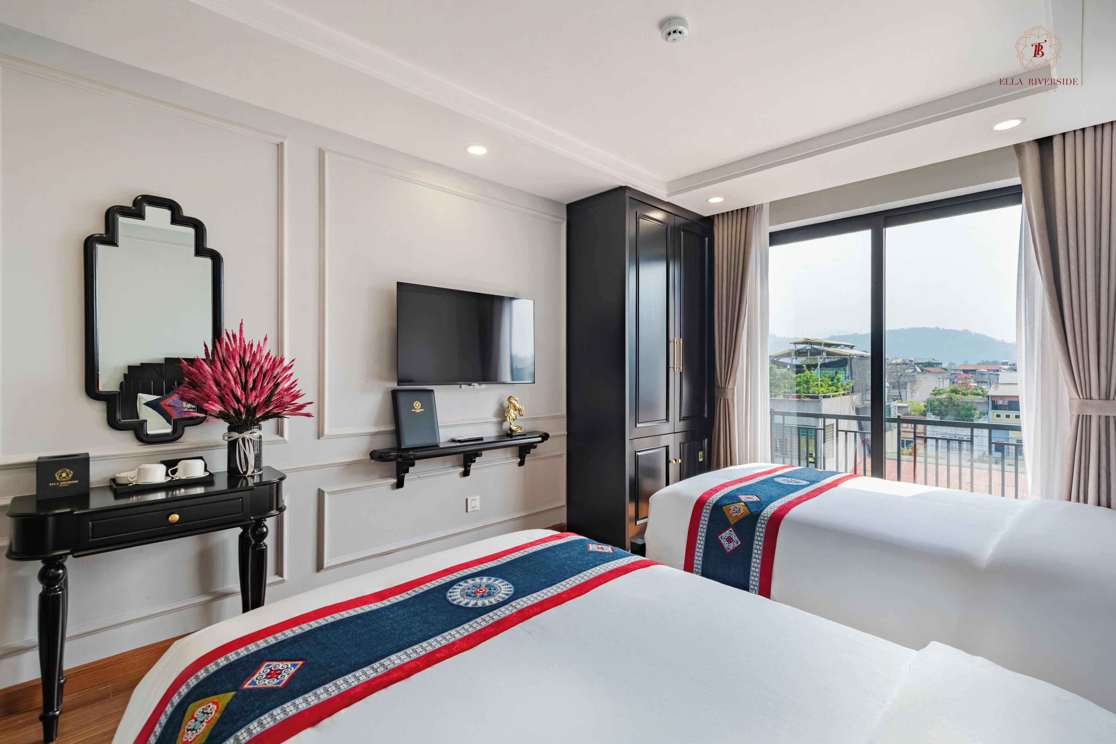 A hotel room with two beds, a TV, a black console table, a mirror, and a view of the city through large windows.