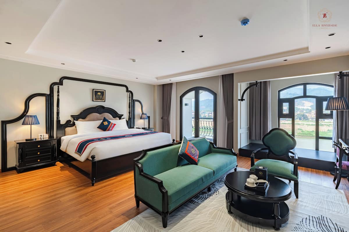 Habitación de hotel con una cama grande, un sofá verde, una mesita y vistas al exterior a través de amplias ventanas.