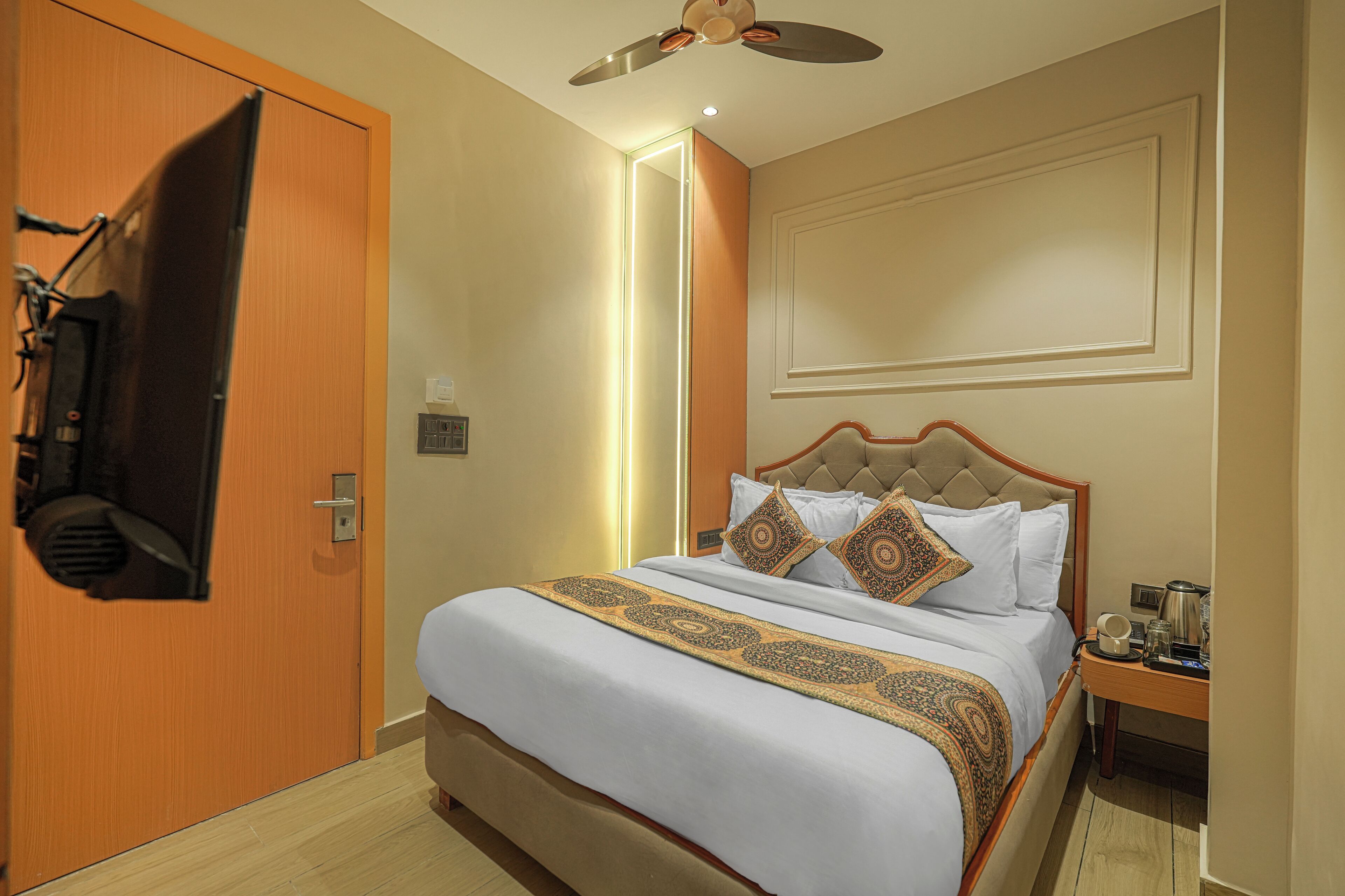 A hotel room with a bed, a wooden door, a ceiling fan, and a nightstand with a coffee maker.