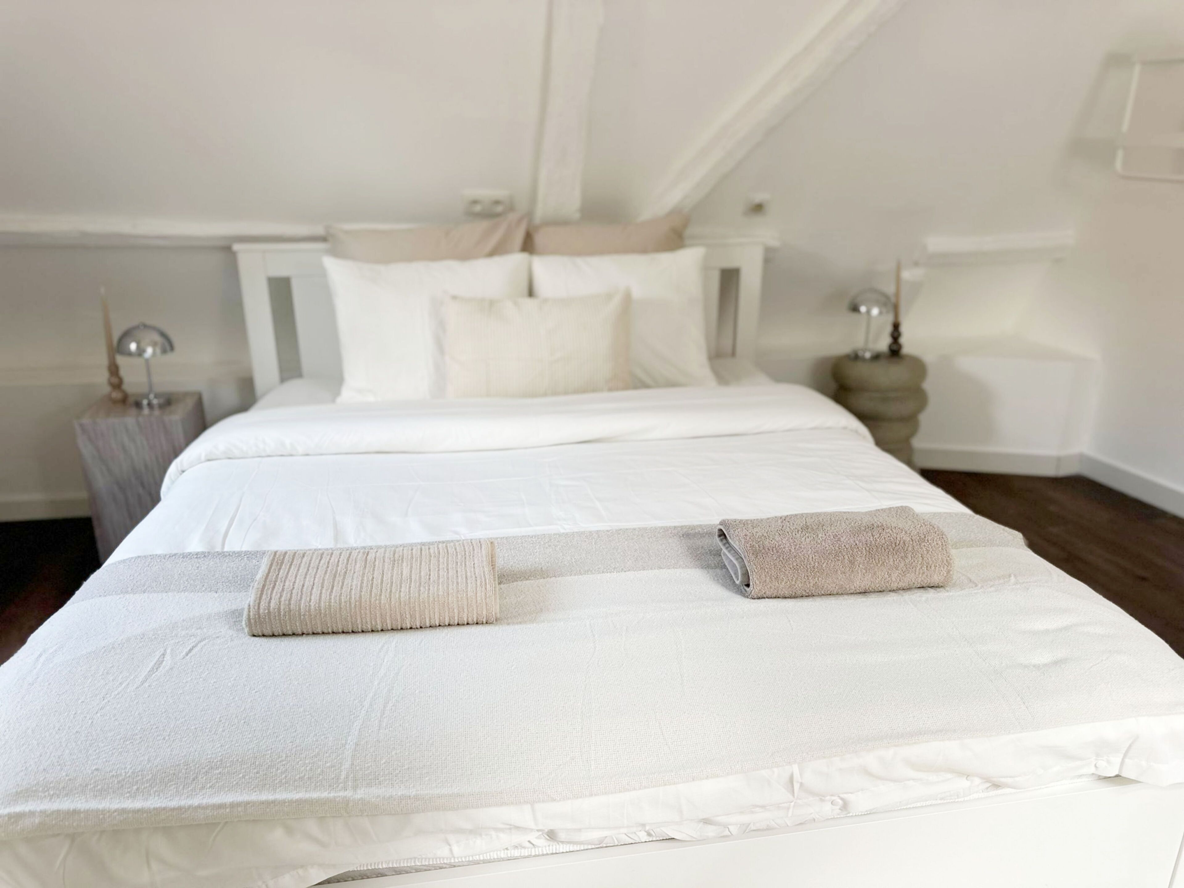 Un lit bien fait, avec du linge de lit blanc et deux serviettes pliées, une table de chevet avec une lampe et une petite table de nuit également avec une lampe.