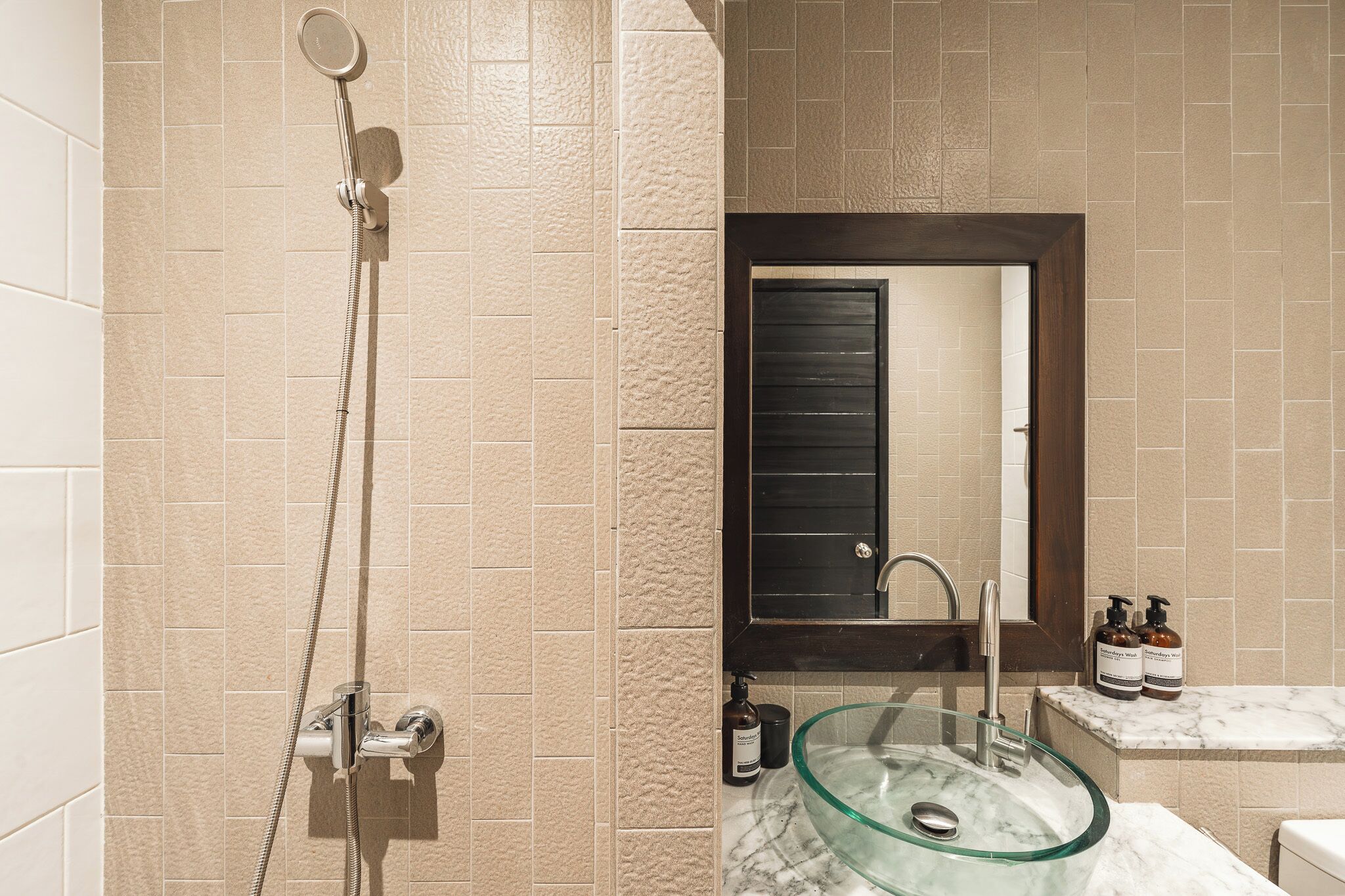 A bathroom with beige tiles, a glass sink, a shower with a handheld showerhead, and a dark-framed mirror.