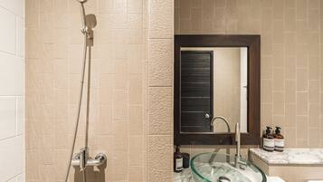 A bathroom with beige tiles, a glass sink, a shower with a handheld showerhead, and a dark-framed mirror.