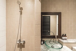 Un baño con azulejos color beige, un lavamanos de vidrio, una ducha con cabezal de ducha de mano y un espejo con marco oscuro.
