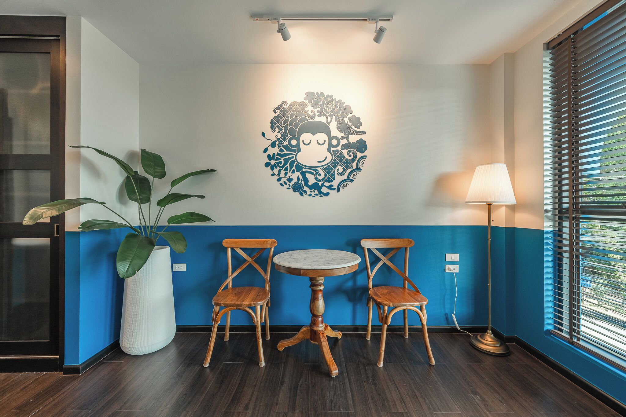 A room with a blue and white wall, a round table with two chairs, a large potted plant, and a wall-mounted artwork of a monkey surrounded by flowers.