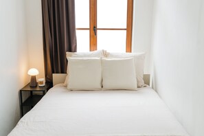 A neatly made bed with white linens and pillows, a bedside table with a lamp and a small container, and a window with curtains. - Modern 3BR Apt for 6 with Patio Ghent (Ghent)