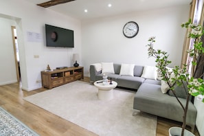 A living room with a grey sofa, a white coffee table, a wooden side table, a TV mounted on the wall, and a clock. - Modern 3BR Apt for 6 with Patio Ghent (Ghent)