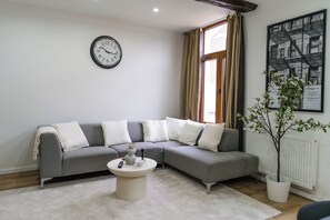 A modern living room with a grey sectional sofa, a round coffee table, a wall clock, a potted plant, and a framed picture on the wall. - Modern 3BR Apt for 6 with Patio Ghent (Ghent)