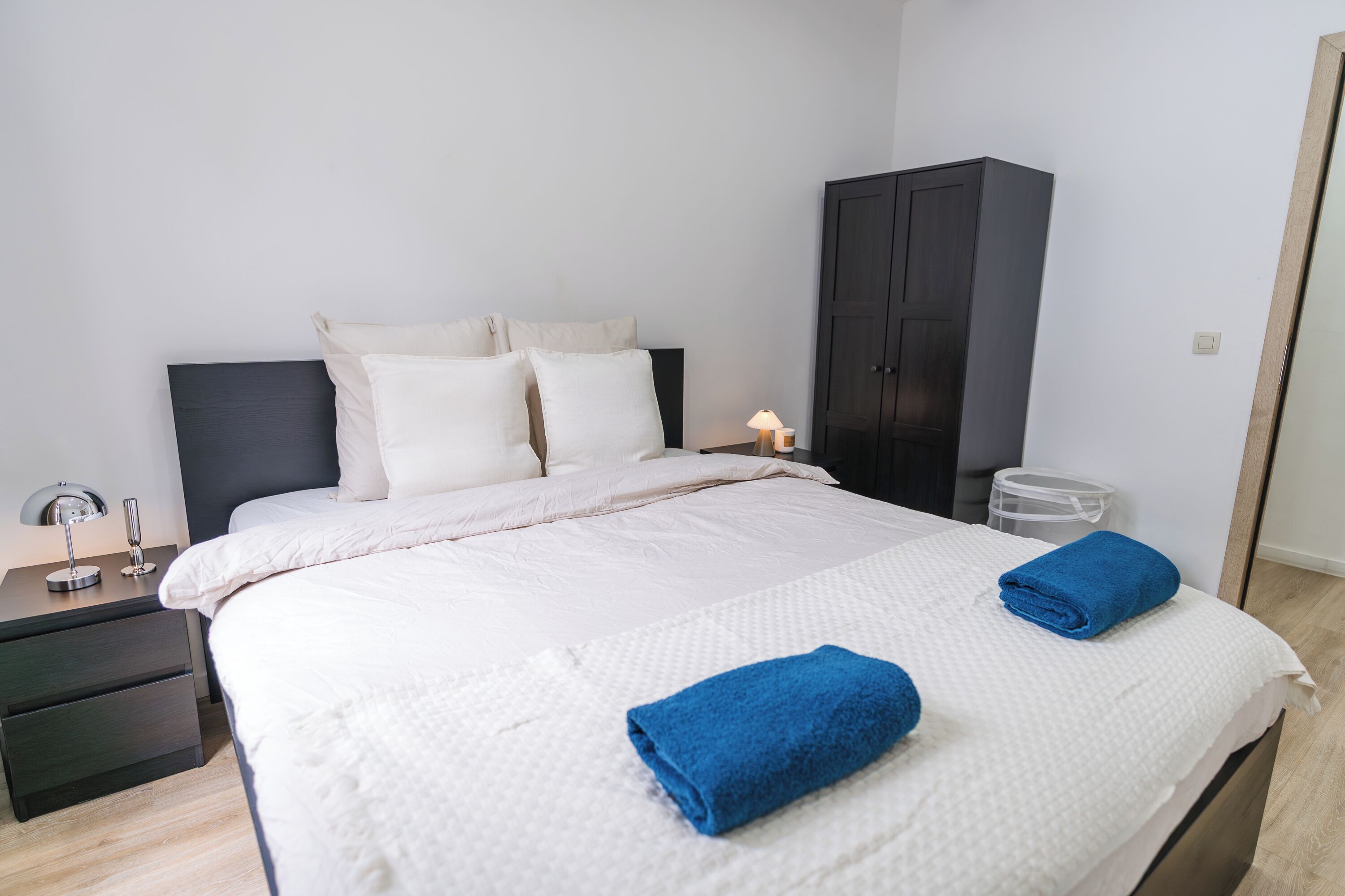 A neatly made bed with white bedding and two blue towels, a dark wooden wardrobe, and a small bedside table with a lamp.