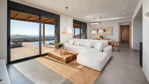 A modern living room with a large sofa, a coffee table, and a dining area with a view of the mountains. - Vista Blue Villa Naxos - 2 (Τοπική Κοινότητα Βίβλου)