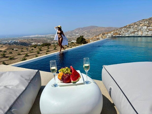A poolside setting with a table set for two, featuring a fruit platter and glasses of white wine.