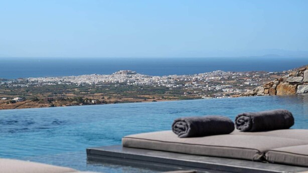 A poolside area with lounge chairs and towels, overlooking a coastal cityscape with a clear view of the sea and distant hills. - Vista Blue Villas Naxos | 16 Guests (Τοπική Κοινότητα Βίβλου)