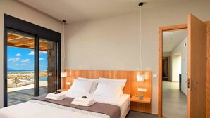 A modern bedroom with a large bed, wooden headboard, bedside tables, and a view of a desert landscape through a glass door. - Vista Blue Villas Naxos | 16 Guests (Τοπική Κοινότητα Βίβλου)