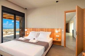 A modern bedroom with a large bed, wooden headboard, bedside tables, and a view of a desert landscape through a glass door. - Vista Blue Villas Naxos | 16 Guests (Τοπική Κοινότητα Βίβλου)