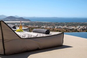A reclining lounge chair with a bottle of wine and grapes, overlooking a coastal landscape with buildings and mountains. - Vista Blue Villa Naxos - 1 (Τοπική Κοινότητα Βίβλου)