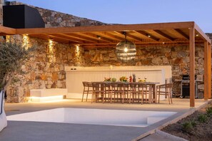An outdoor dining area with a stone wall backdrop, wooden pergola, and a hanging light fixture. - Vista Blue Villa Naxos - 1 (Τοπική Κοινότητα Βίβλου)