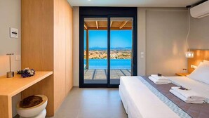 Una habitación de hotel moderna con una cama, un escritorio de madera, un baño pequeño y un cerramiento de vidrio corredera que da a una piscina y vistas panorámicas.
