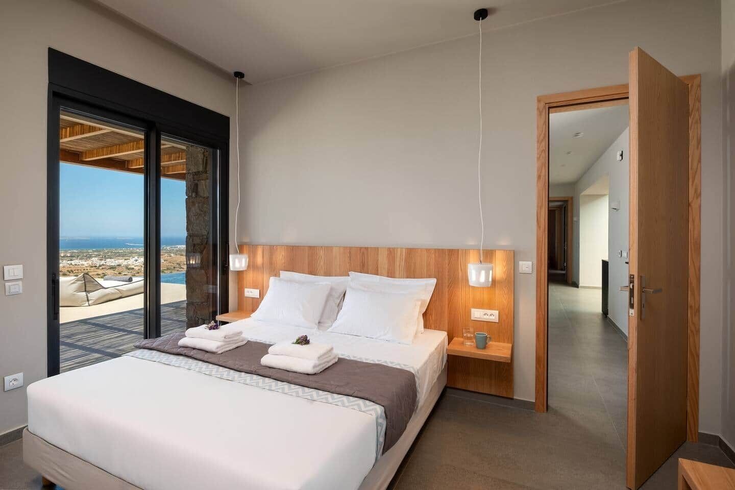 Een moderne slaapkamer met een groot bed, een houten hoofdbord, nachtlampjes en uitzicht op een kustlandschap.