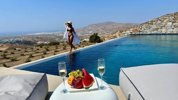 Escena junto a la piscina con una mesa preparada para dos personas, con una bandeja de frutas y dos copas de vino blanco.