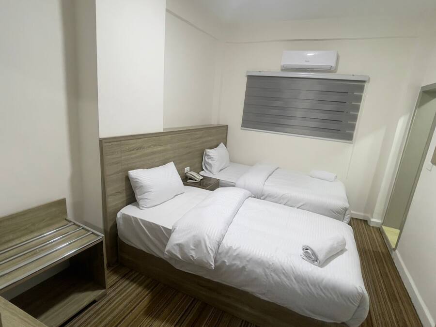A hotel room with two beds, a wooden bench, a bedside table, a telephone, and a wall-mounted air conditioner.