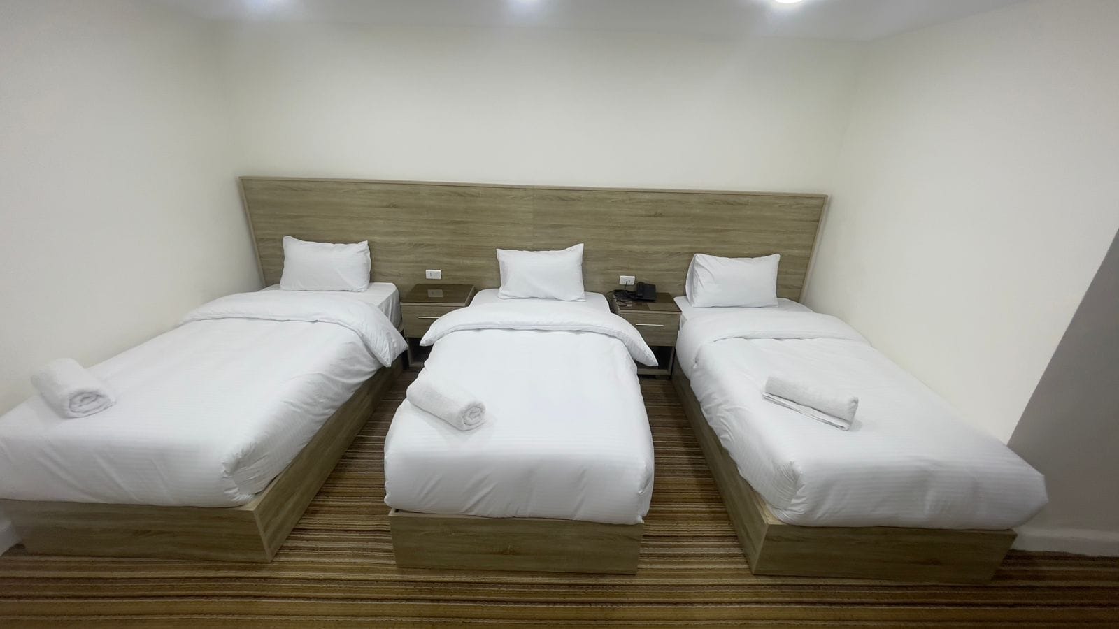 A hotel room with three single beds, wooden headboards, white linens, and a wooden floor.