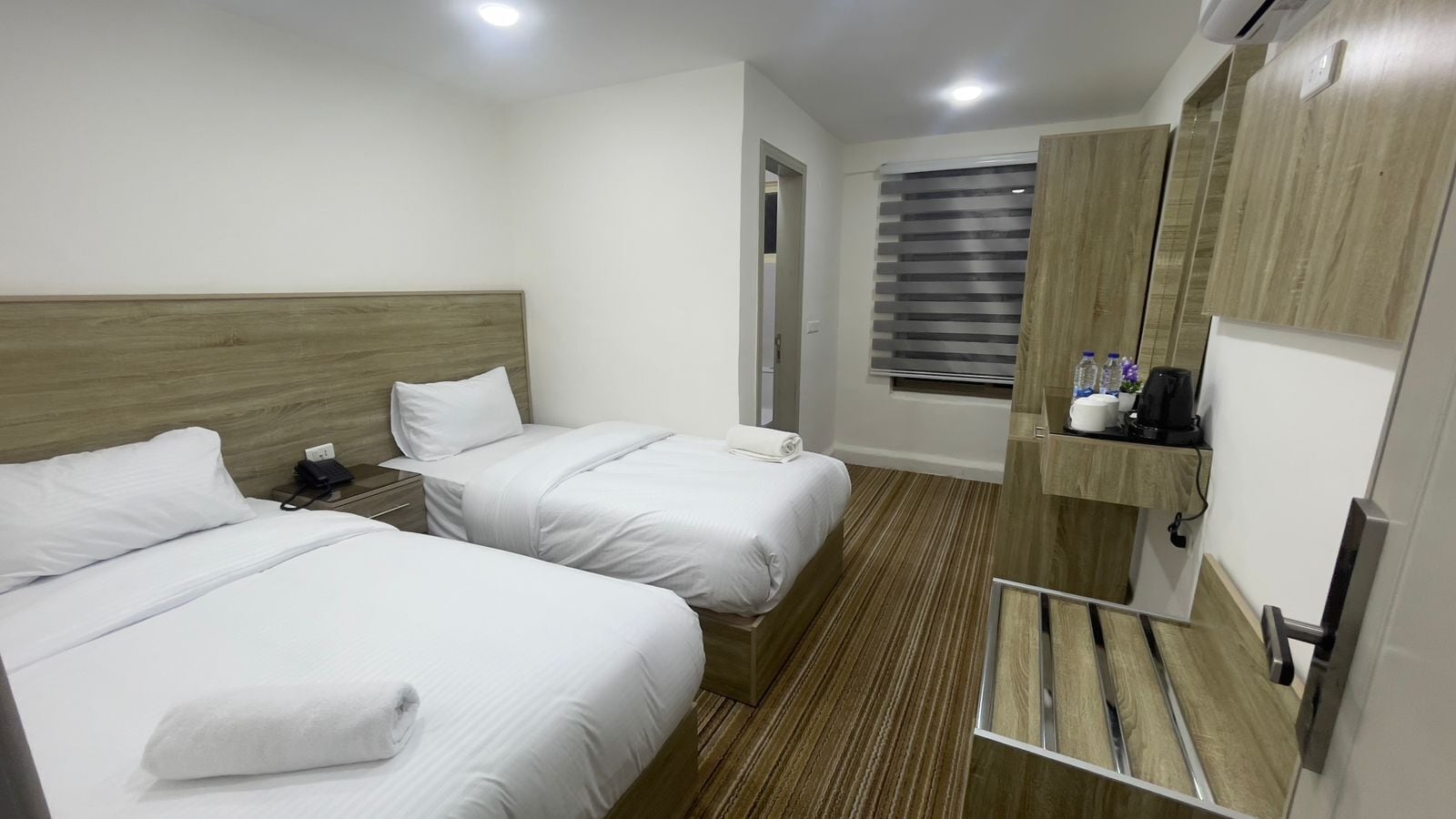 A hotel room with two beds, a wooden headboard, a small bedside table with a phone, a window with blinds, and a built-in shelf with toiletries.