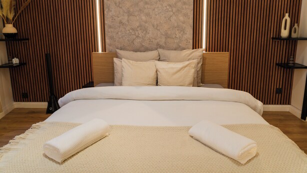 A neatly made bed with a textured white bedspread and two rolled towels, a wooden headboard, and a wall with vertical wooden slats. - Cozy 5BR Apt in the Heart of Ghent (Ghent)