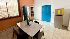 A small, well-lit room with a marble table and chairs, a refrigerator, a blue door, and a window with blinds. - Colonial Home with Private Terrace and Jacuzzi (Santo Domingo)