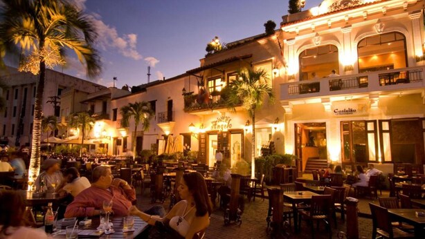 A bustling outdoor dining area with tables and chairs, palm trees, and a building with a sign reading "Castelo." - Colonial Home with Private Terrace and Jacuzzi (Santo Domingo)