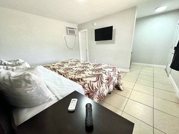 Una camera d'albergo con un letto, una TV fissata al muro, un telecomando su un tavolo e una porta che dà su un'altra stanza.