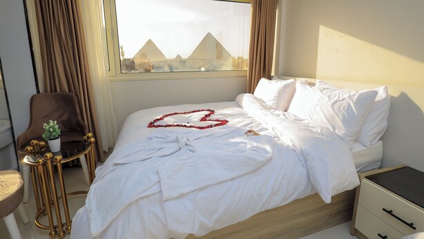 Een hotelkamer met een bed, een nachtkastje, een stoel, een kleintje tafeltje met een plant en uitzicht op piramides door het raam.