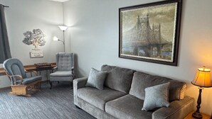 Ein Wohnzimmer mit grauem Sofa, blauem Sessel, einem Holztischchen, einer Stehlampe und einem Bilderrahmen mit einem Stadtbild.