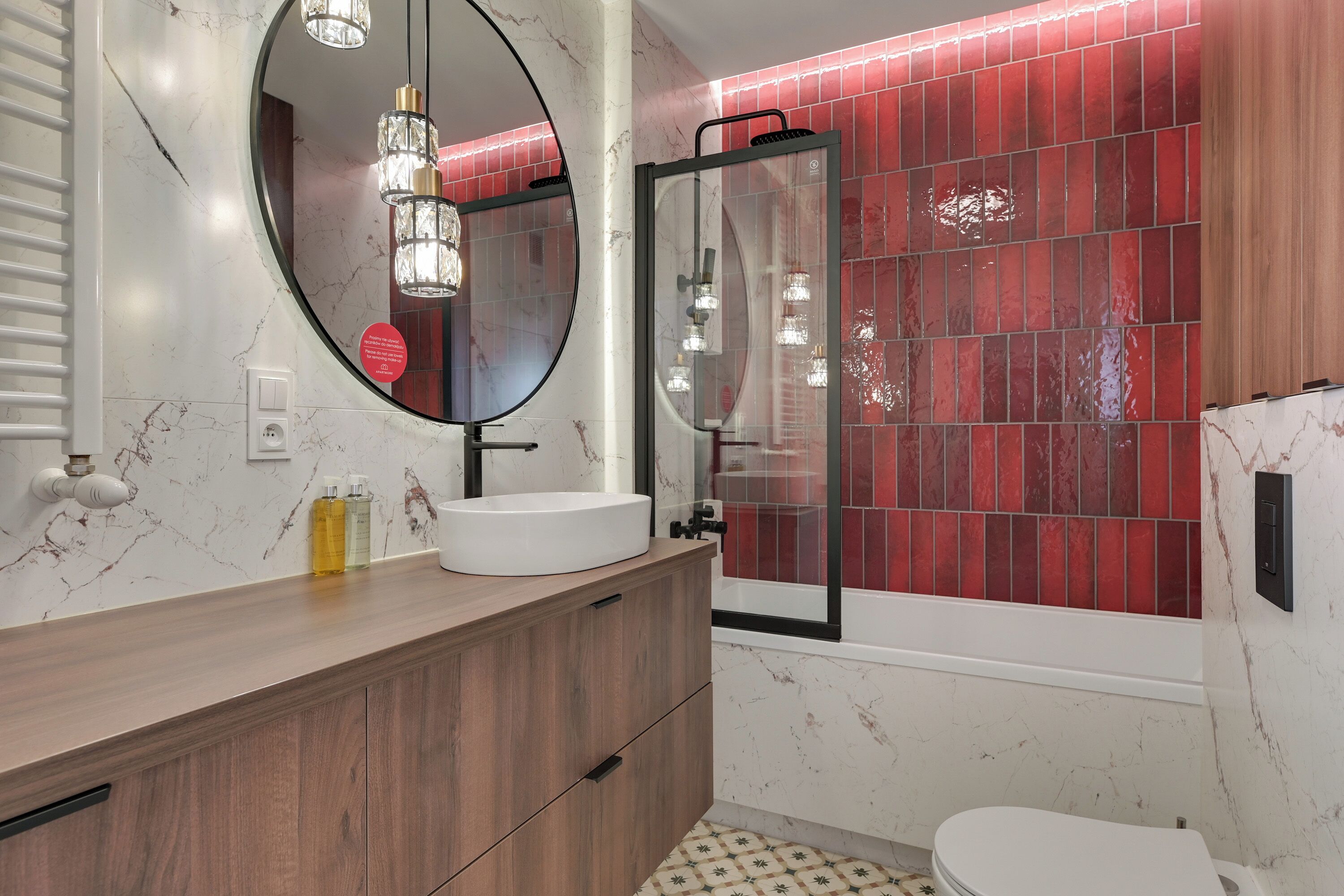 A bathroom with a round mirror, a wooden vanity, a white sink, a glass shower enclosure, and red-tiled walls.