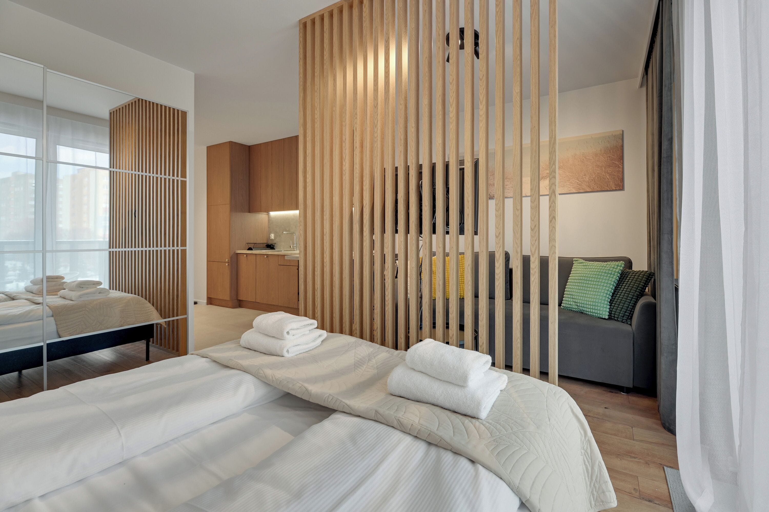 A modern hotel room with a large bed, a sofa, a wooden partition, and a view of the city.