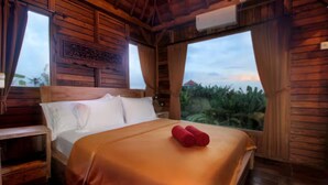 A wooden room with a bed, a nightstand, and a window overlooking greenery. - Tri Datu Palace (Sukawati)