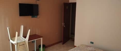 Una habitación con cama, una silla, una mesita, televisor montado en la pared y una puerta que da a otra estancia.