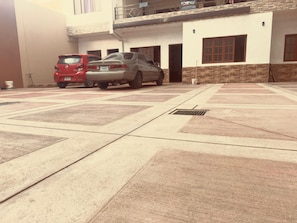 Un espace de stationnement où sont garées deux voitures : un SUV rouge et une berline argentée, devant un immeuble à la façade en briques et doté de nombreuses fenêtres.