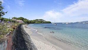 Una playa con agua cristalina, barcos y casas al fondo.