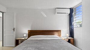 Un dormitorio con cabecera de madera, una cama con manta gris, dos mesitas de noche con lámparas y un equipo de aire acondicionado en la pared.