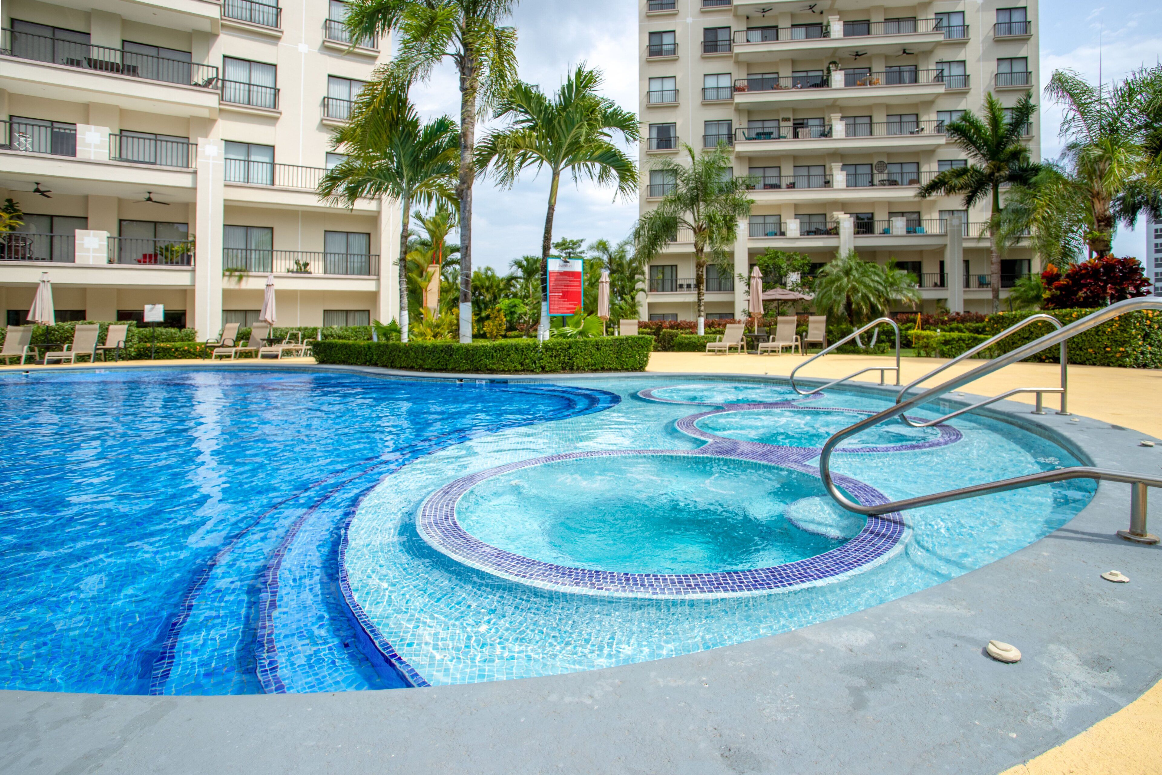 Una piscina con borde de azulejos, rodeada de palmeras y un edificio de hotel de varios pisos.