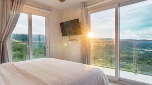Ein Schlafzimmer mit einem großen Bett, einem an der Wand montierten Fernseher und einem Balkon mit Blick auf Grünflächen und Hügel.