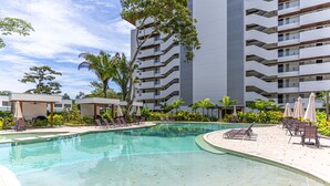 A swimming pool with lounge chairs and umbrellas, surrounded by a multi-story building with balconies. - Selva Coral 0701 (Jacó)