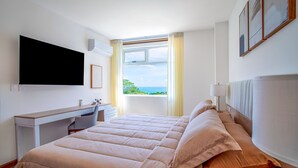 A bedroom with a bed, a desk, a TV, and a window with a view of the ocean. - Selva Coral 0701 (Jacó)