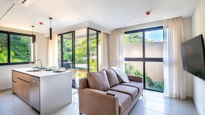A modern living space with a kitchen island, a sofa, and large windows offering a view of greenery. - Torres del Mar 1202 (Jacó)