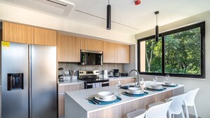 A modern kitchen with stainless steel appliances, wooden cabinets, and a large window offering a view of trees. - Torres del Mar 1202 (Jacó)