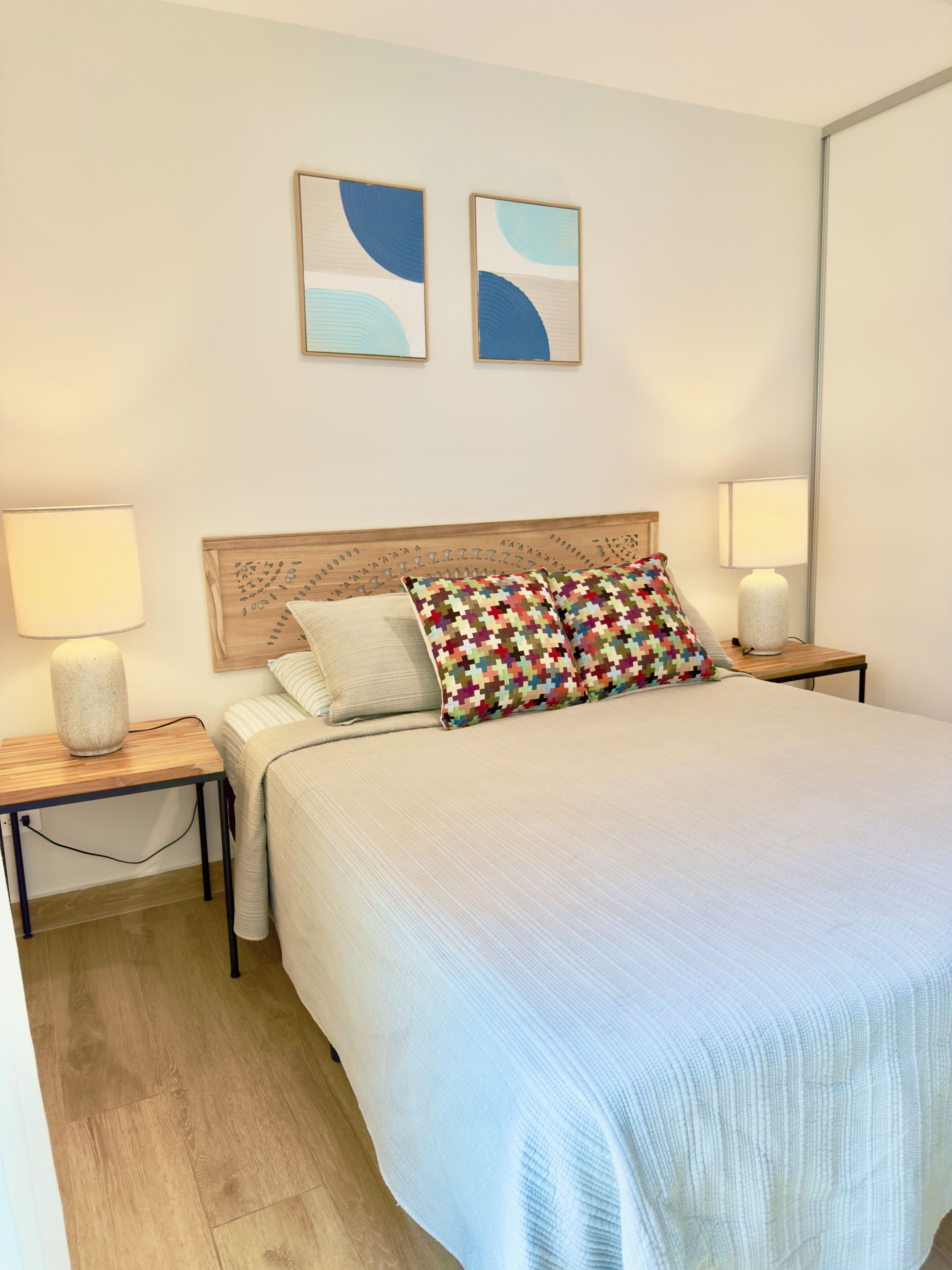 A neatly made bed with a patterned pillow, two wall-mounted abstract paintings, and bedside lamps on wooden nightstands.