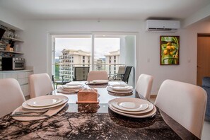 Un coin repas avec une table dressée pour quatre personnes, une vue sur les bâtiments à travers une porte vitrée et un appareil de climatisation fixé au mur.