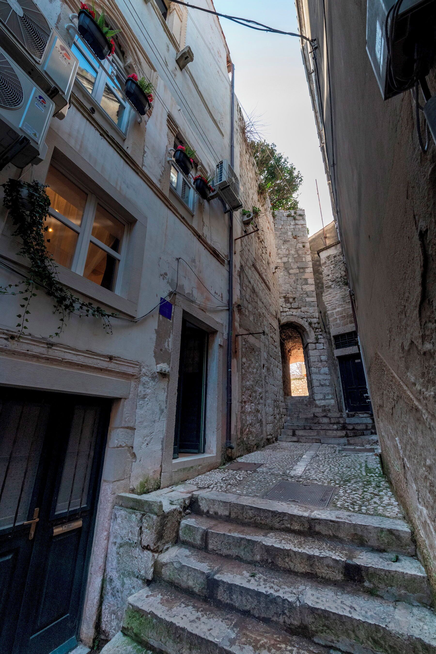 A narrow stone staircase leading up to a building with multiple windows, some with plants.