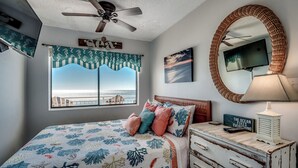 Een slaapkamer met een bed, een plafondventilator, een groot raam met uitzicht, een dressoir, een spiegel en een tv aan de muur.