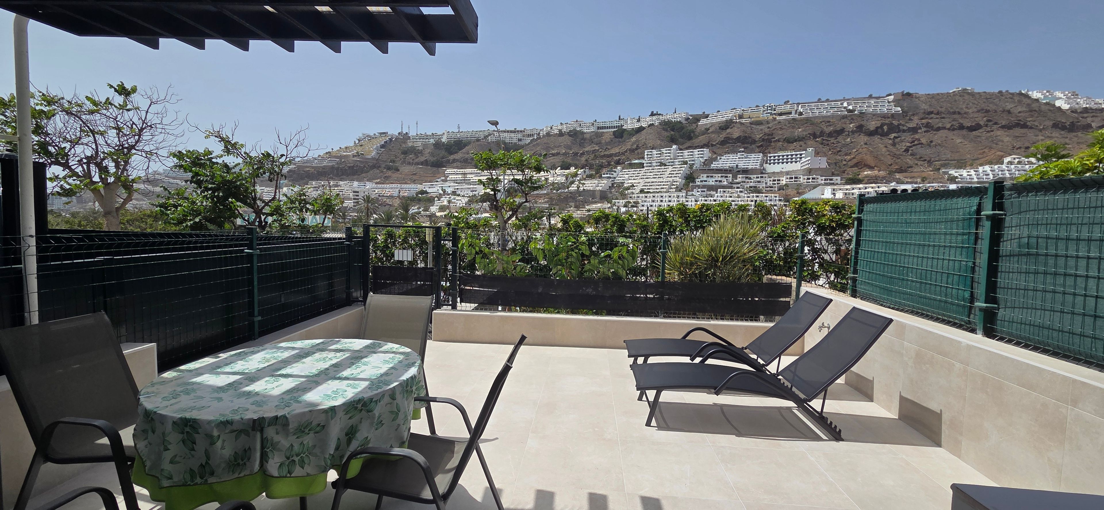 Un patio al aire libre con una mesa y sillas, vista a una colina con edificios y una cerca verde.